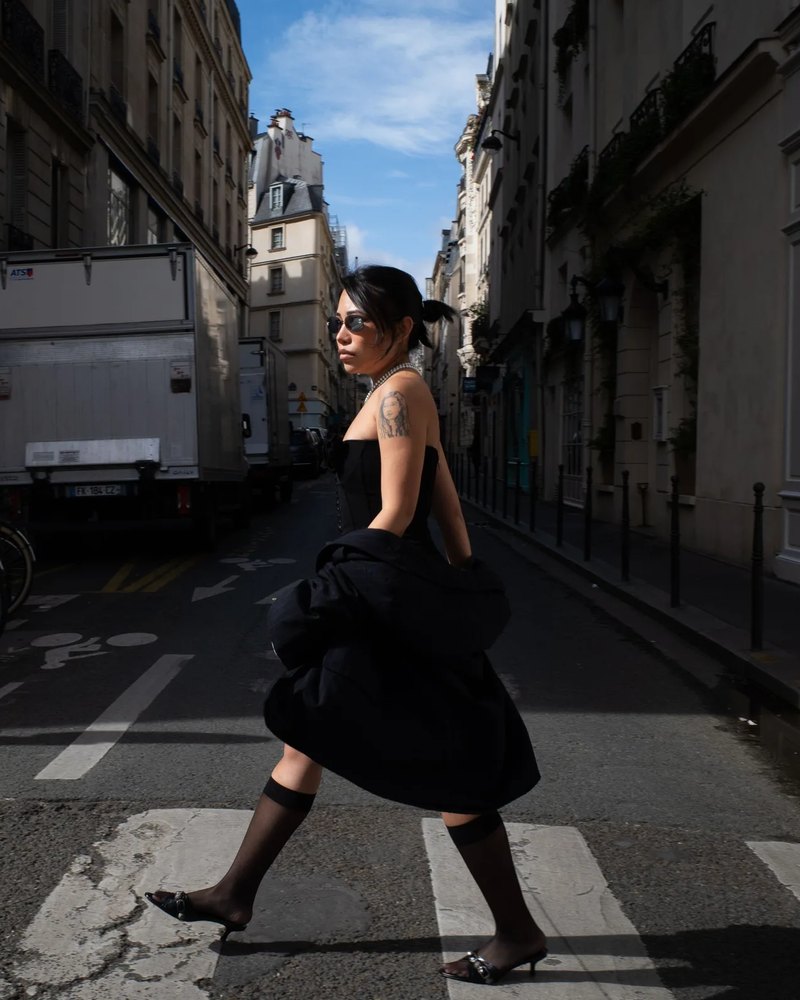 Femme en robe bustier noire, veste noire au bras, chaussettes voile et mules, traversant une rue de Paris
